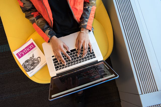 Woman in a yellow chair sits at a laptop with a programming book beside her