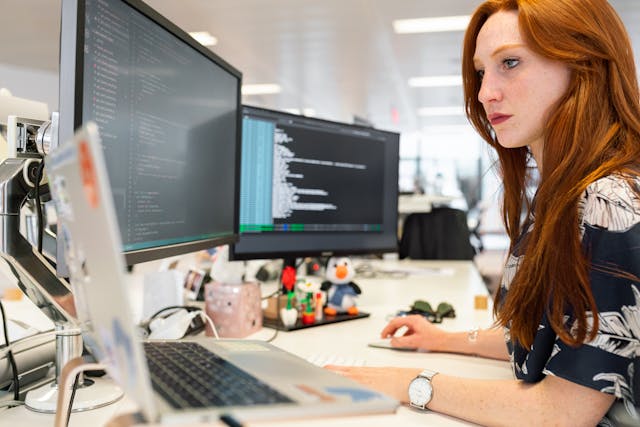 woman with red hair sits in front of multiple computer screens