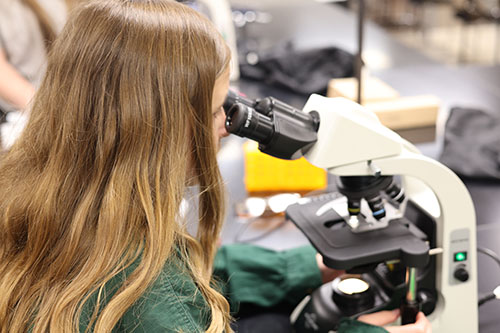 Young woman with long brown hair wearing a long-sleeve green shirt looks into a microscope