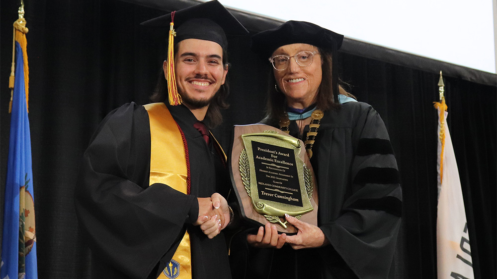 Two people in graduation robes shake hands while also holding a plaque