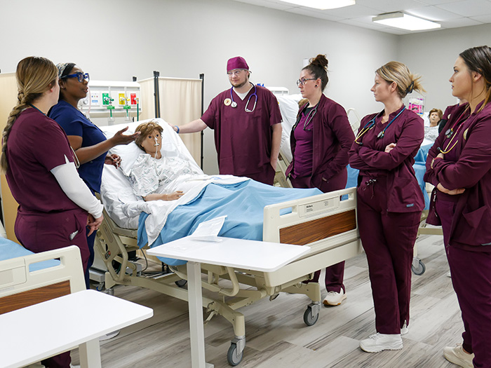 Nurses in maroon scrubs listen to a nurse in blue scrubs talk, while all are surrounding a simulation mannequin