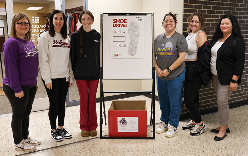 Six women stand along side an easel with a shoe drive poster on it, and a red box underneath it