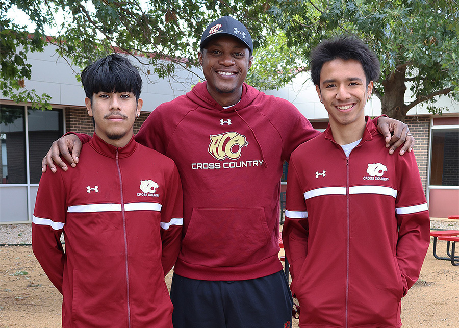 Three men in maroon and gold jackets stand together in front of some trees