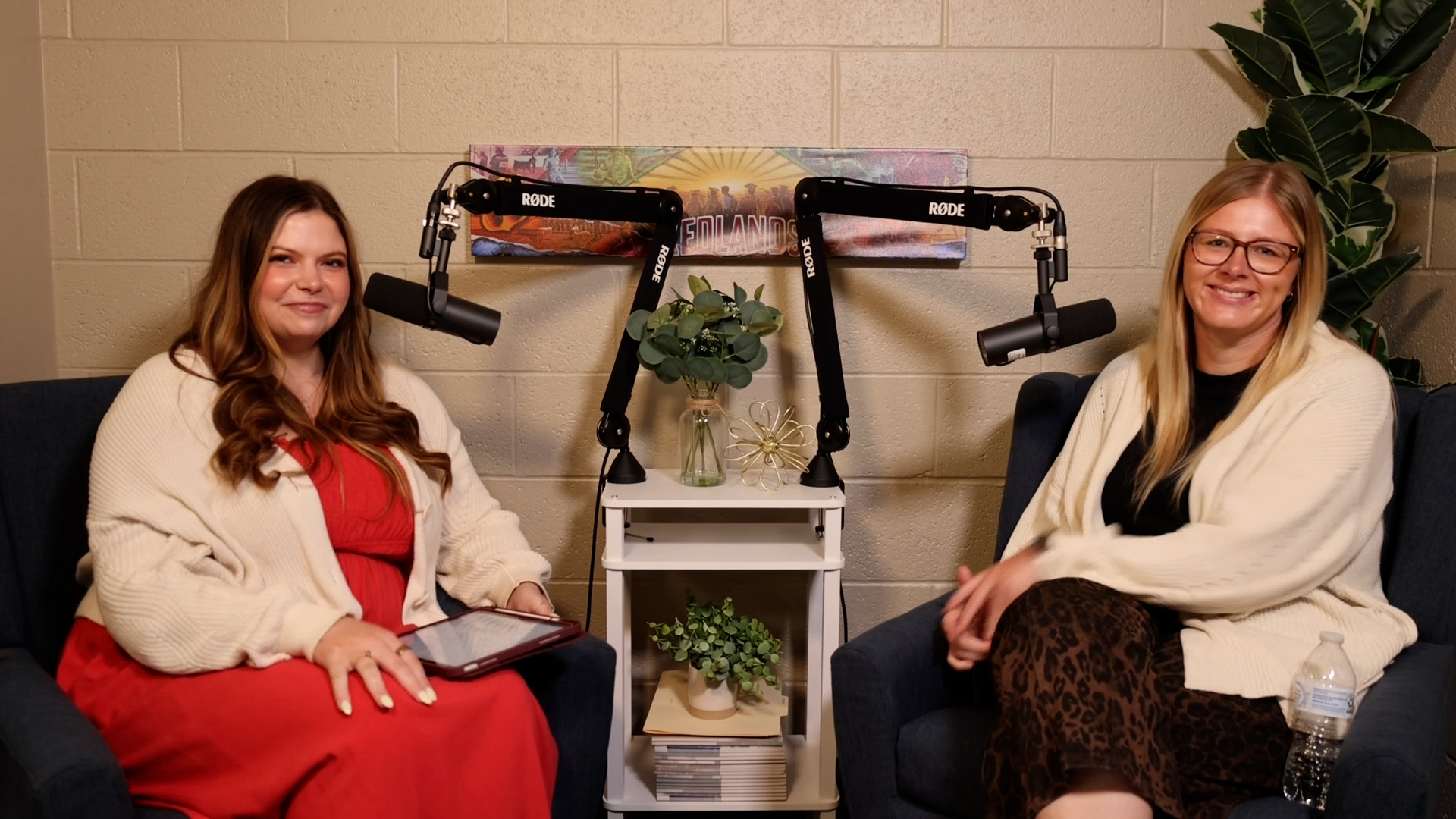 Woman in orange dress and sweater sits next to woman in cheetah print pants while talking in microphones