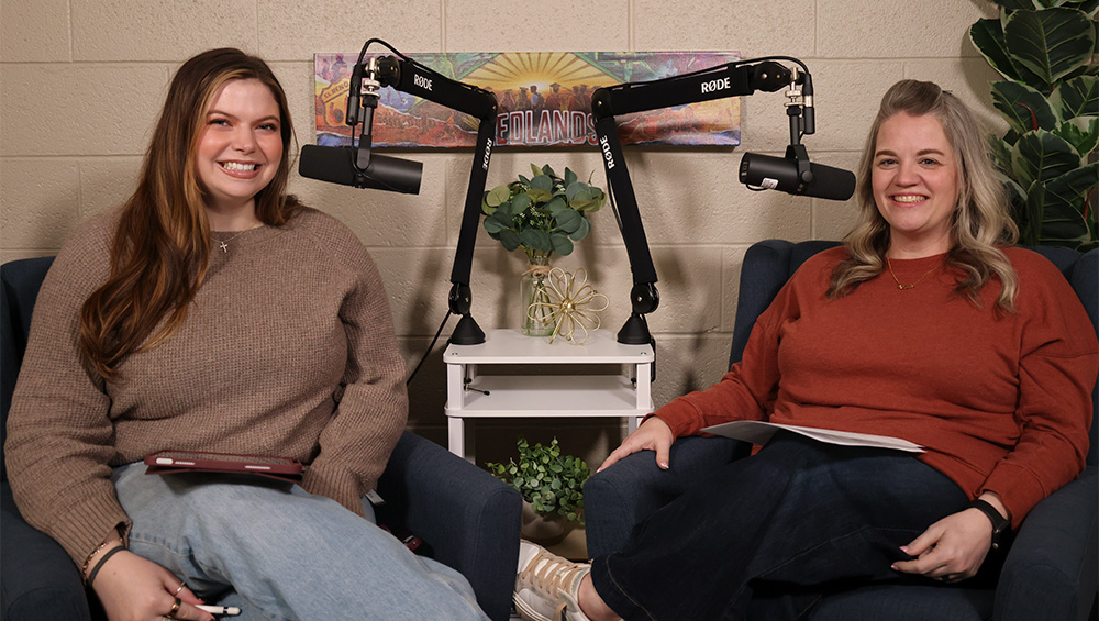 Woman in gray sweater smiles next to woman in red sweater while sitting at podcast microphones