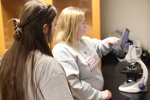 Student looks on as a staff member assists with setting up a microscope